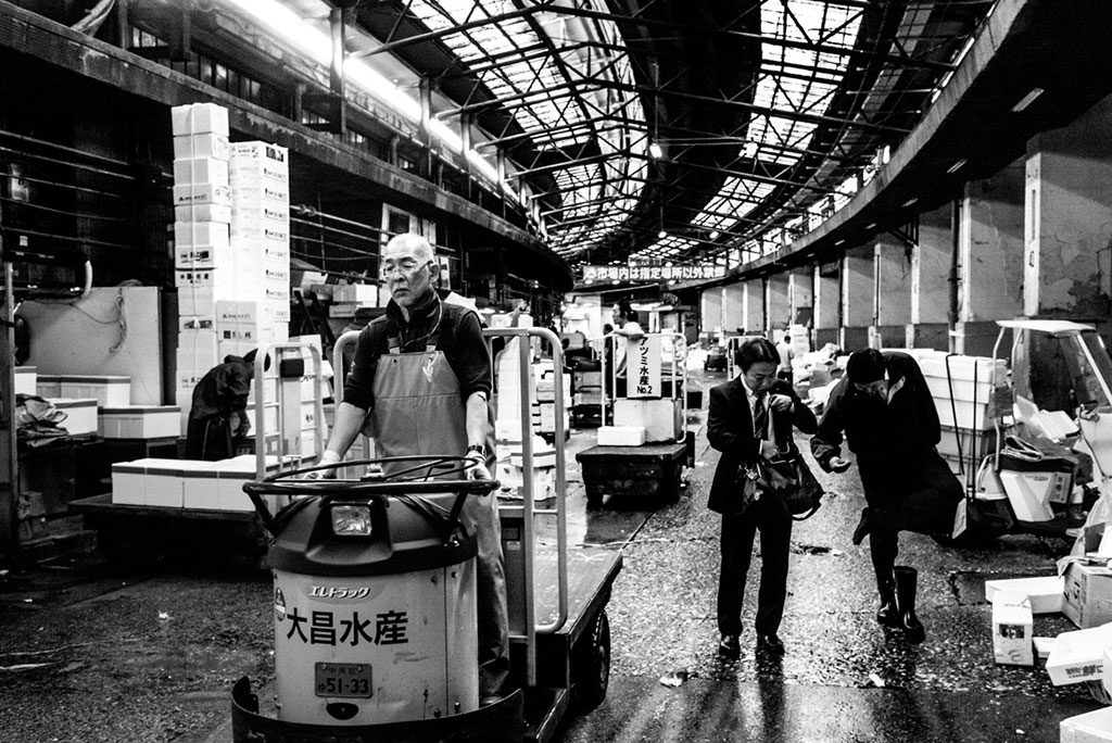 Black and white photo from Tokyo Tsukiji photo book. A man wearing a suit rummages in his bag, another takes off his shoe and checks his foot. An old worker drives a cart.